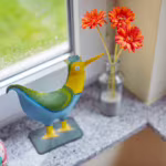 Das Bild zeigt einen bunten Metallvogel und eine Blumenvase auf einer Fensterbank.