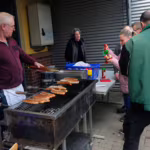 Das Bild zeigt den Grill und Besucher, die Bratwurst kaufen.